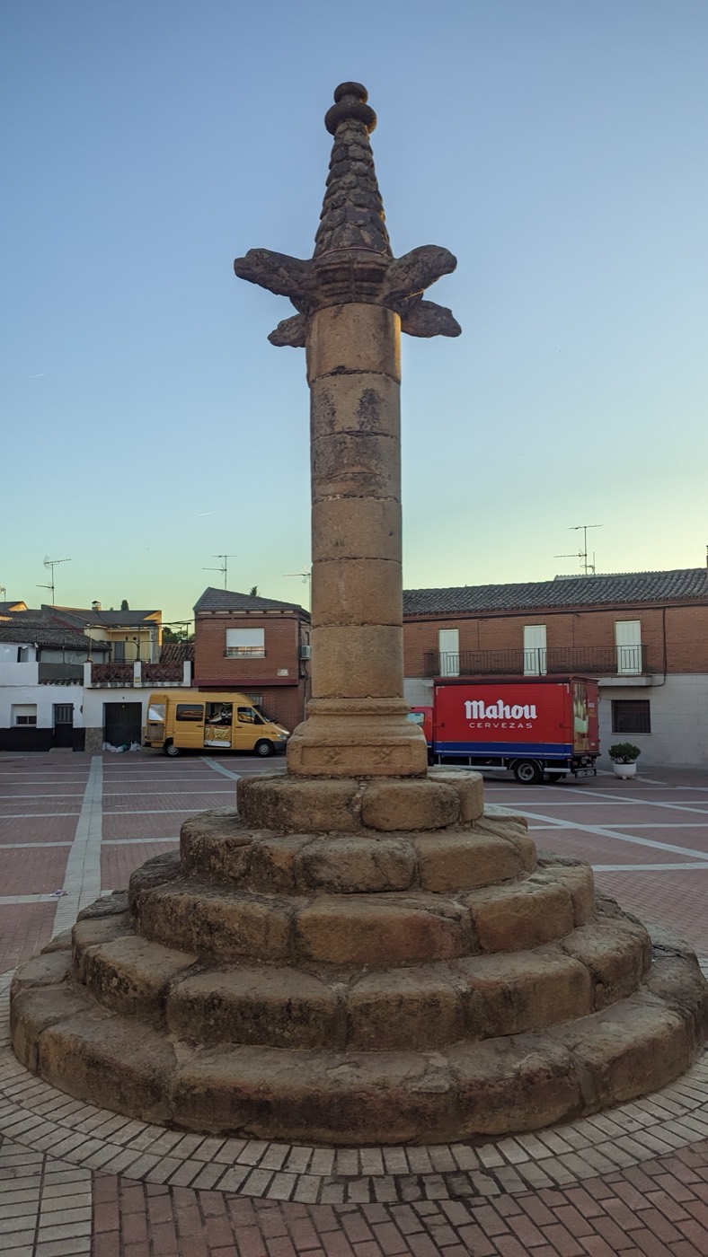 El rollo de San Román de los Montes en la plaza, al atardecer: cinco escalones redondos, una columna de granito y, arriba, cuatro cabezas de perro y un copete de piedra.